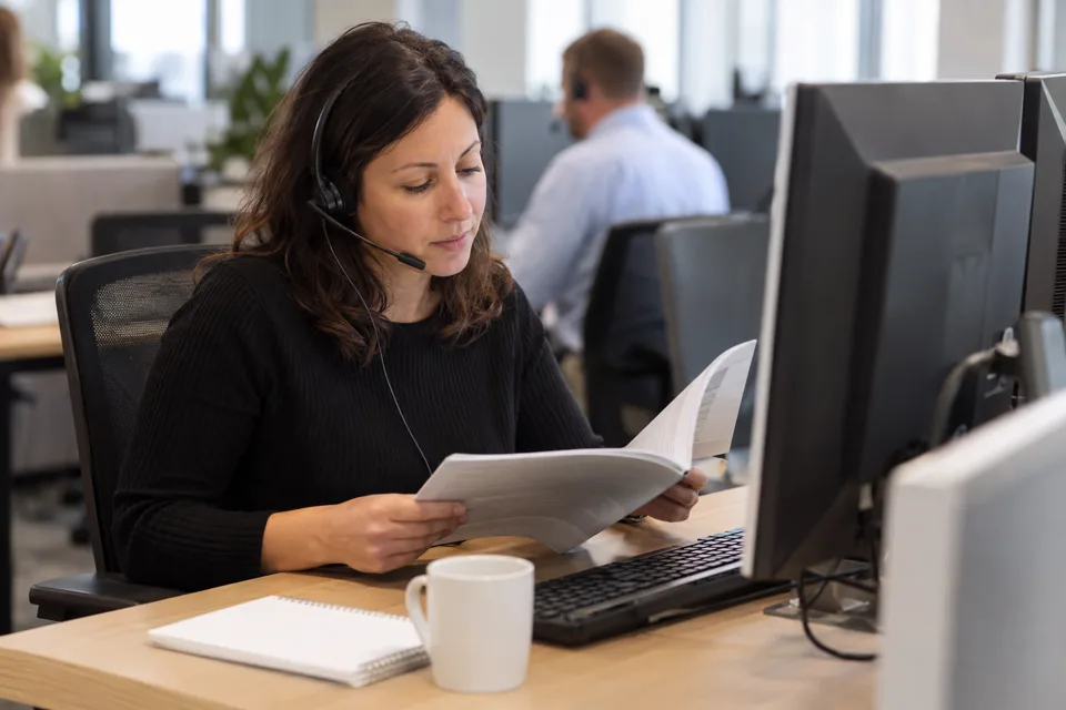A customer support specialist on a headset reads an open instruction booklet at her desk in an open-plan office.